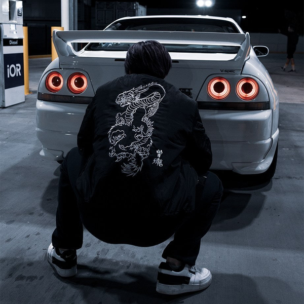 Japanese retro fashion brand dragon embroidered jacket men's women's autumn winter bomber jacket work wear casual. View of male model back to camera crouching down facing the back of while car.